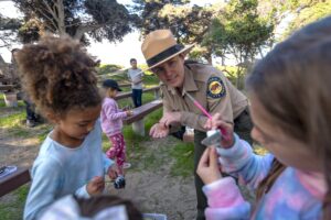 Photograph of an outdoor scene at Pismo State Beach featuring rustic benches and trees featuring children holding arts and crafts projects and a woman dressed in a California State Parks uniform