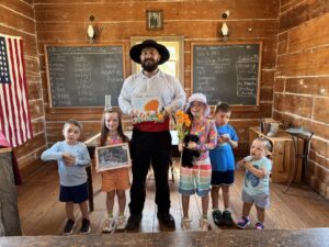 A man dressed in historical attire stands with five children in an old-fashioned schoolroom at Old Town San Diego State Historic Park, holding a book titled Wake Sleepy One as the children hold flowers, drawings, and toys.
