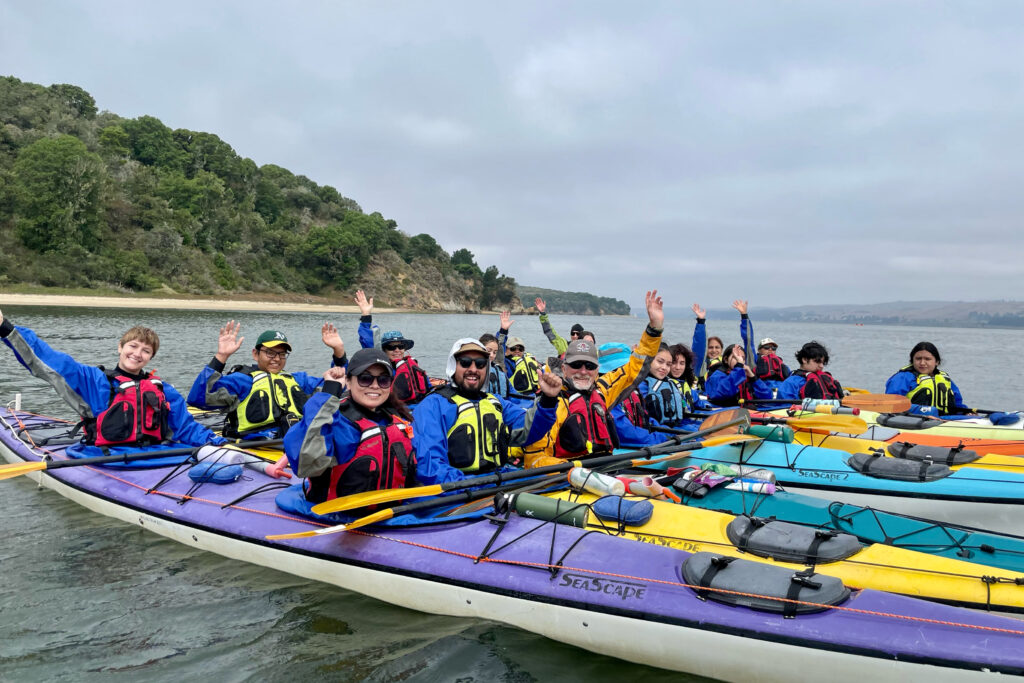 Adults and kids wearing life jackets are sitting in kayaks in Tomales Bay and waving to the camera.