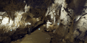 A screenshot of the virtual tour of Mitchell Caverns. It shows the lit interior of the caverns, with a paved path, with rocky walls and stalactites.