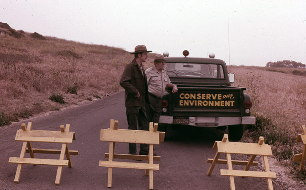 c. 1970 photograph of a road through a grassy hillside; the road is blocked by three wooden barriers behind which stand two men in Parks uniforms leaning against a green pickup truck with the tailgate emblazoned