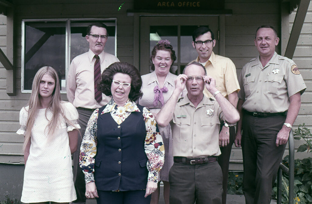 Photographic portrait of seven women and men variously dressed in early 1970s era office attire and Parks uniforms, all standing in two rows on the front porch of a building clad in wooden clapboard siding