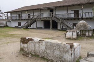 Photograph of a crumbling adobe wall and an adobe horno oven in the middle of a decomposed granite courtyard framed by a two story adobe building.