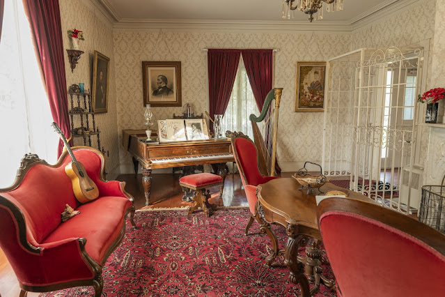 Living Room, Vallejo Home, Sonoma SHP Photograph of a 19th-century-appointed sitting room featuring wood and red velvet upholstered furniture, musical instruments, and formal portraiture.