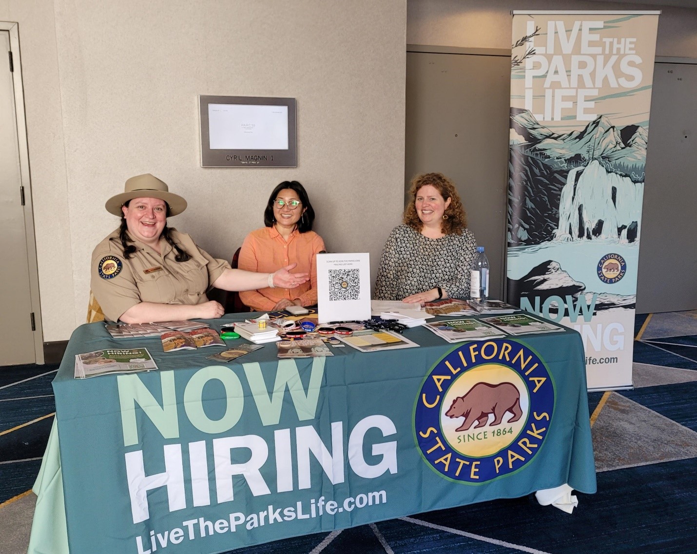 LGBTQ+ Career Table Three women variously waring business casual attire and California State Parks uniforms are seated behind an outreach event table; both the table cloth and an adjacent stand-up banner are emblazoned with the California State Parks logo and the words, "NOW HIRING LiveTheParksLife.com"