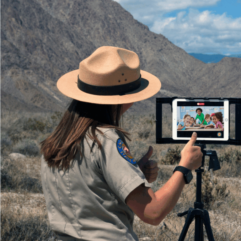 Photograph of a California State Park Interpreter in uniform standing in an arid landscape blooming with wildflowers facing away from the camera and gesturing towards a tablet device on a tripod displaying five children gathered around a table.