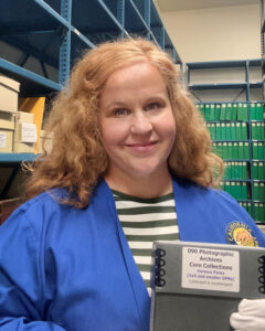 Portrait of Rebecca Crowther, standing in front of metal shelving, holding a paper archival documents box, wearing a blue smock and white cotton gloves