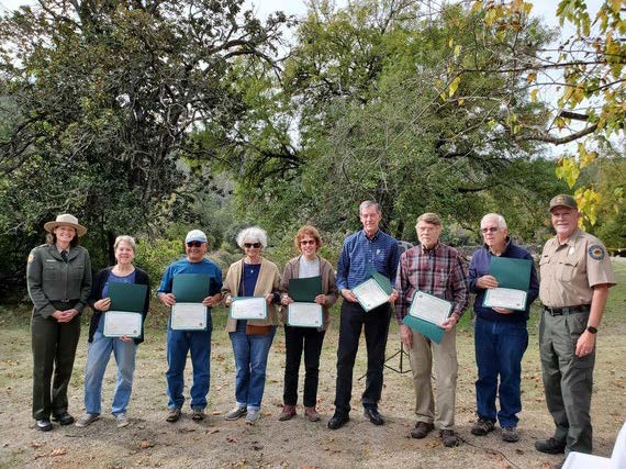 Sierra Gold Sector Poppy Awardees Judy Beck, Andy Berg, Robert Coats, Tad Kitada, Mary Ann Coleman, Marsha Lewis, and George Sanford with staff members, Rob Griffith and Jean Rhyne. Photo by Jeremy Lin. A group of volunteers stand in a line holding certificates while flanked by uniformed State Park employees; the group are arranged in a line and standing outdoors in front of a stand of trees.
