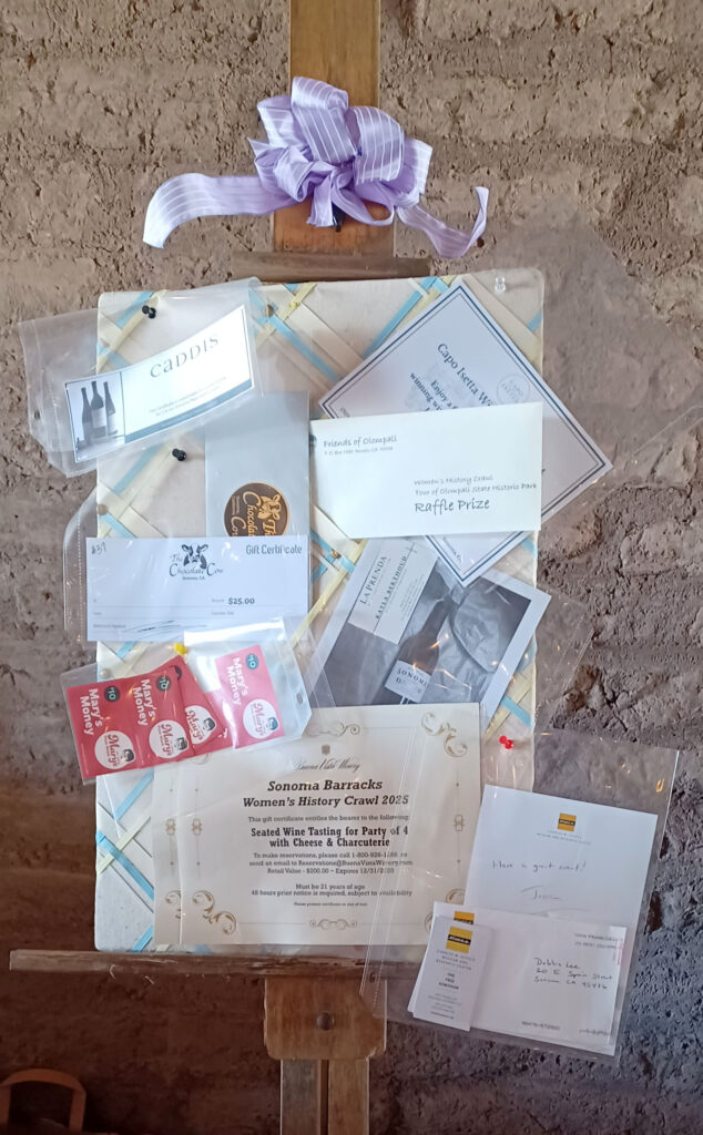 Photgraph of a freestanding wooden easel infront of an adobe wall; the easel is holding a canvas decorated with ribbons and is holding up several sheets of paper pinned to it, each representing a prize donated by a partner organization.