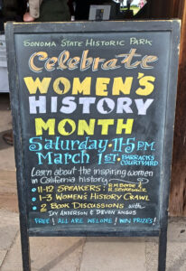 Photograph of a freestanding chalkboard sign reading "Sonoma State Historic Park, Celebrate Women's History Month, Saturday 11-5 P.M. March 1st Barracks Courtyard, Learn about the inspiring women in California history, 11-12 Speakers: R. M. Beebe & R. Senkewicz, 1-3 Women's History Crawl, 2 Book Discussions with Ivy Anderson & Devon Angus, Free! All Are Welcome! Win Prizes!"