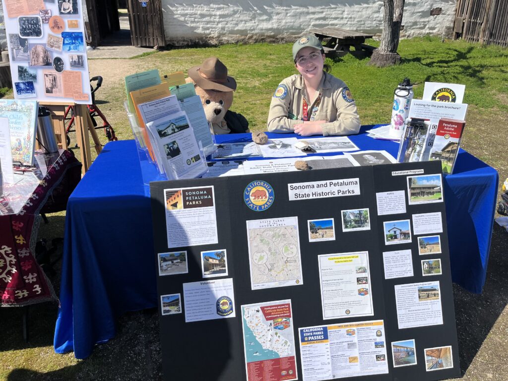 Photograph of a woman in a Calfironai State Parks Park Aide Uniform sitting behind a table liberally festooned with informational pamphlets, outreach materials, and interpretive items.