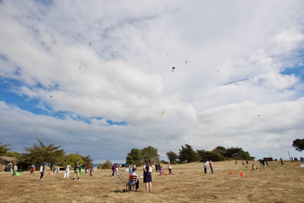 People flying kites in an open grassy field under a partly cloudy sky at Candlestick Point State Recreation Area during a community Kite Day event. Groups of children and adults are scattered across the area, with colorful kites visible in the sky above. Orange cones and trees border the field.