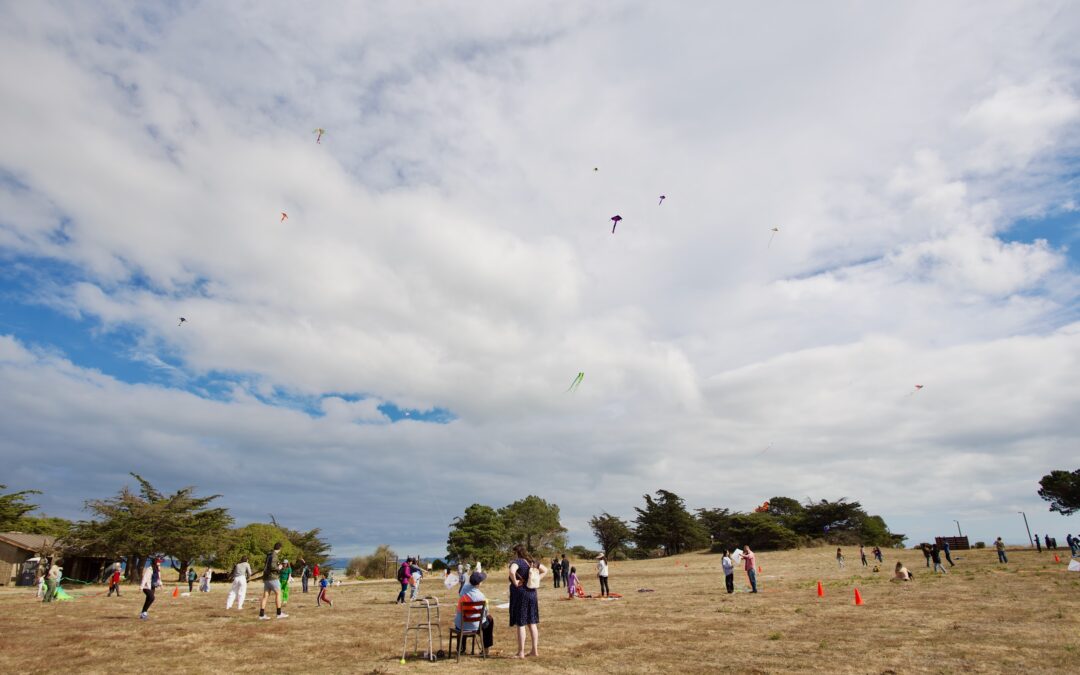 People flying kites in an open grassy field under a partly cloudy sky at Candlestick Point State Recreation Area during a community Kite Day event. Groups of children and adults are scattered across the area, with colorful kites visible in the sky above. Orange cones and trees border the field.