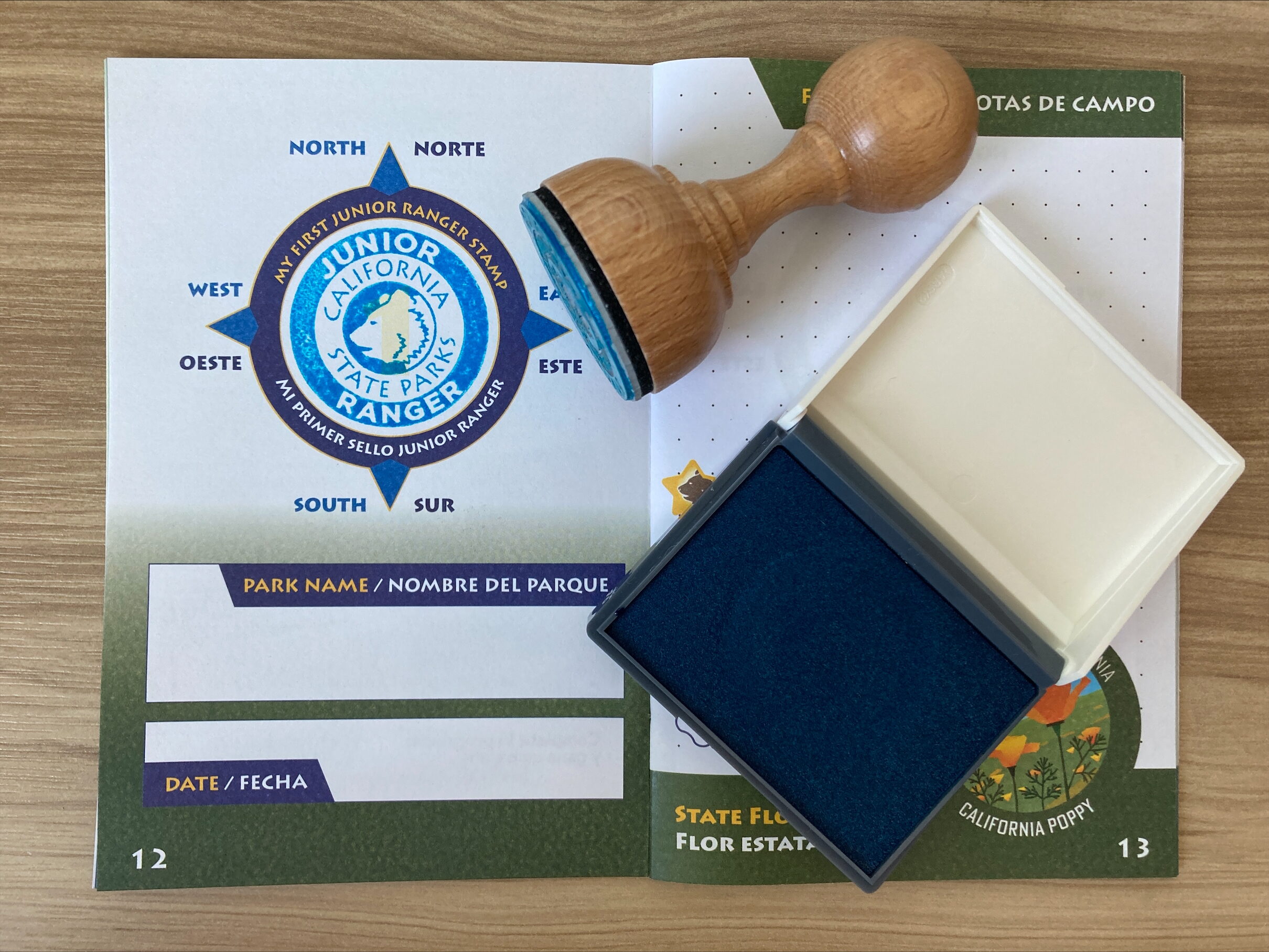 Photograph of an open booklet with a stamp and inkpad resting on top of it; the open booklet page has been stamped with ink in the shape of a bear head and text reading "JUNIOR RANGER CALIFORNIA STATE PARKS)."
