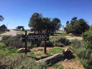 A wooden sign marks the entrance to Carpinteria State Beach, surrounded by greenery and trees under a clear blue sky, with the ocean in the background