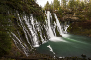 A stunning view of Burney Falls in McArthur-Burney Falls Memorial State Park, with cascading water flowing over a moss-covered cliff into a turquoise pool surrounded by forest.