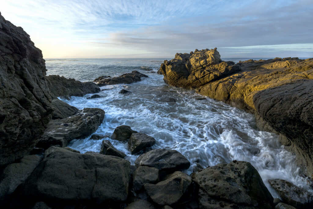 Waves crash against rugged coastal rocks at Leo Carrillo State Park under a soft blue sky during early morning light.