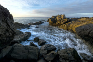 Waves crash against rugged coastal rocks at Leo Carrillo State Park under a soft blue sky during early morning light.