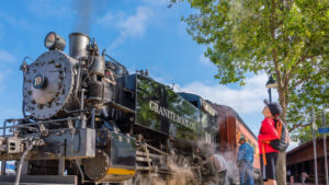 A child in a red shirt and cap looks up at the historic Granite Rock Co. #10 steam locomotive at the California State Railroad Museum. The black engine is emitting steam, and two adults stand nearby. The scene is set on a sunny day with blue skies and green trees.