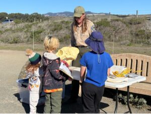 A park interpreter at Hearst San Simeon State Park shows three children a replica elephant seal skull. The kids, holding notebooks, listen attentively around a table with educational items like rope and models.