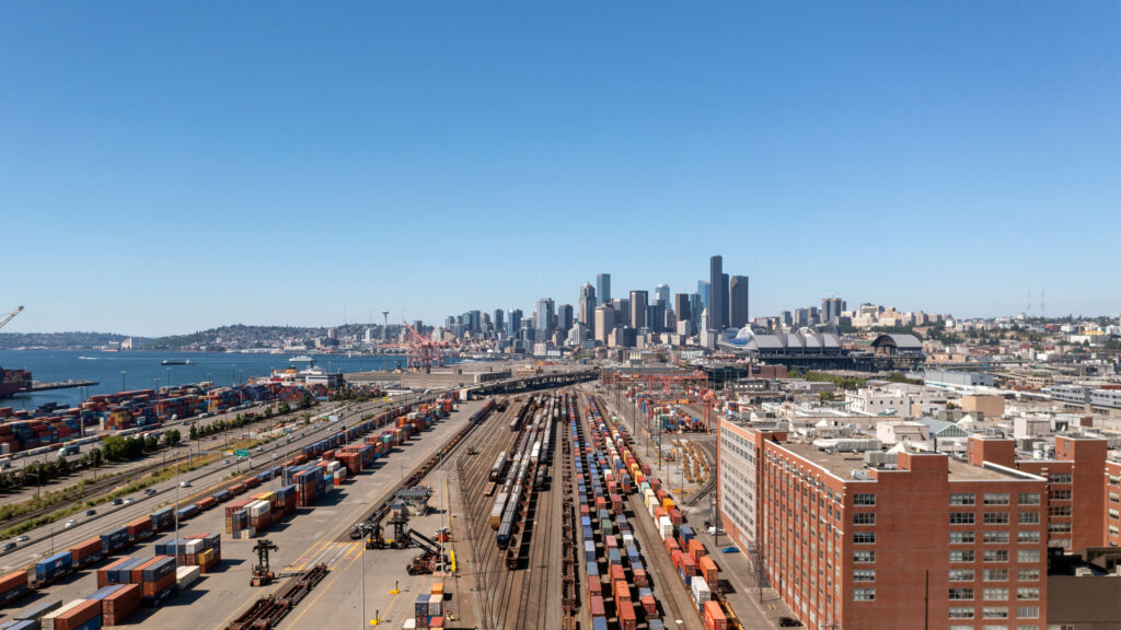 A wide aerial view of Seattle, Washington, showing the downtown skyline with tall buildings under a clear blue sky. In the foreground, there is a busy freight yard with numerous train tracks and colorful shipping containers