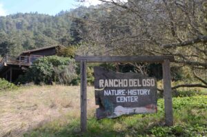 A rustic wooden sign reading "Rancho Del Oso Nature-History Center" stands in a grassy clearing in front of a rustic building, surrounded by trees and forested hills.