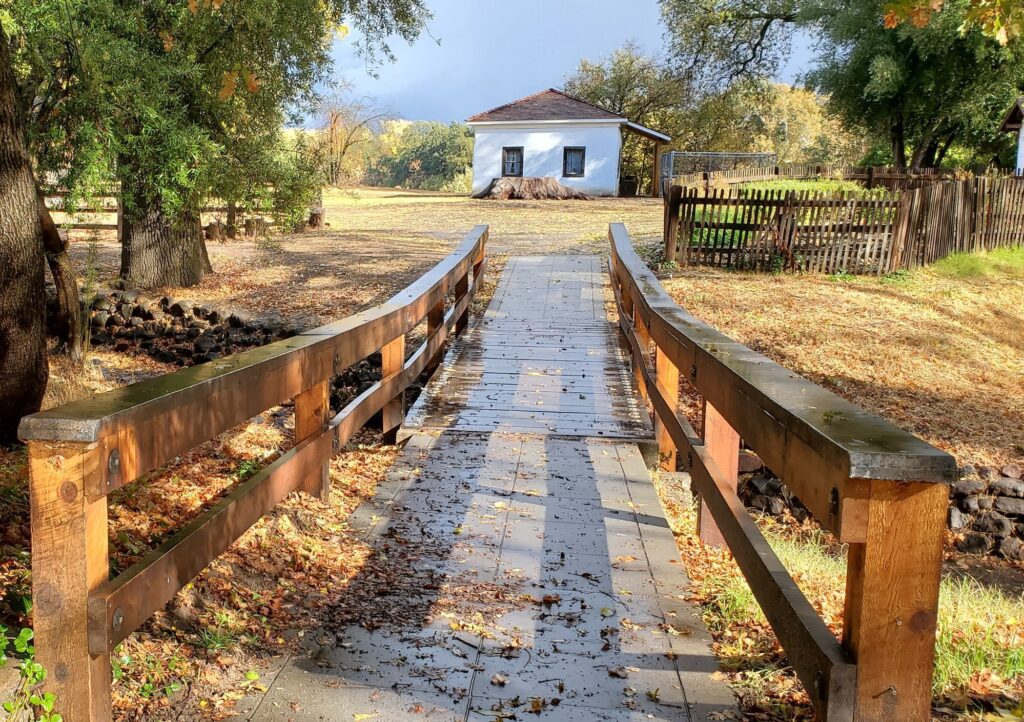 A wooden footbridge leads to a small white adobe building at William B. Ide Adobe State Historic Park, surrounded by trees and a rustic wooden fence.