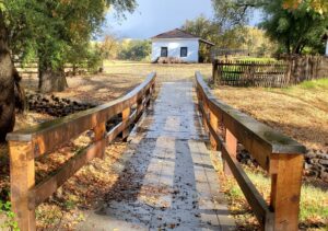 A wooden footbridge leads to a small white adobe building at William B. Ide Adobe State Historic Park, surrounded by trees and a rustic wooden fence.