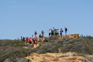 A group of hikers stands and walks along a trail on a coastal bluff beneath a clear sky.