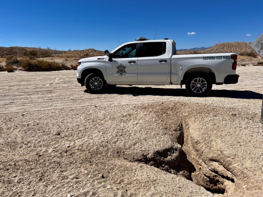 Photograph of a white California State Parks law enforcement pickup truck parked on a sandy desert road; in the foreground is an irregular depression in the ground, and in the background are low hills and distant mountains.