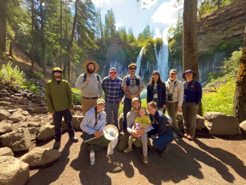 Photograph of 10 adults and 1 child standing in front of a tall waterfall and several tall conifer trees.