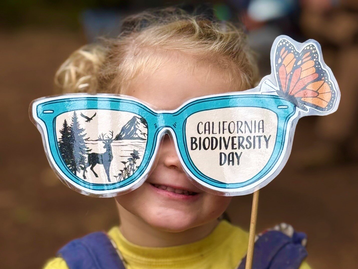 CA Biodiversity Day 2 Photograph of a child holding up a mask obscuring their eyes; the mask is in the shape of glasses and features an illustration of a woodland scene in one lens, the words "Calfiroani biodiversity Day" in the other lens, and an illustration of a Monarch Butterfly atop it.