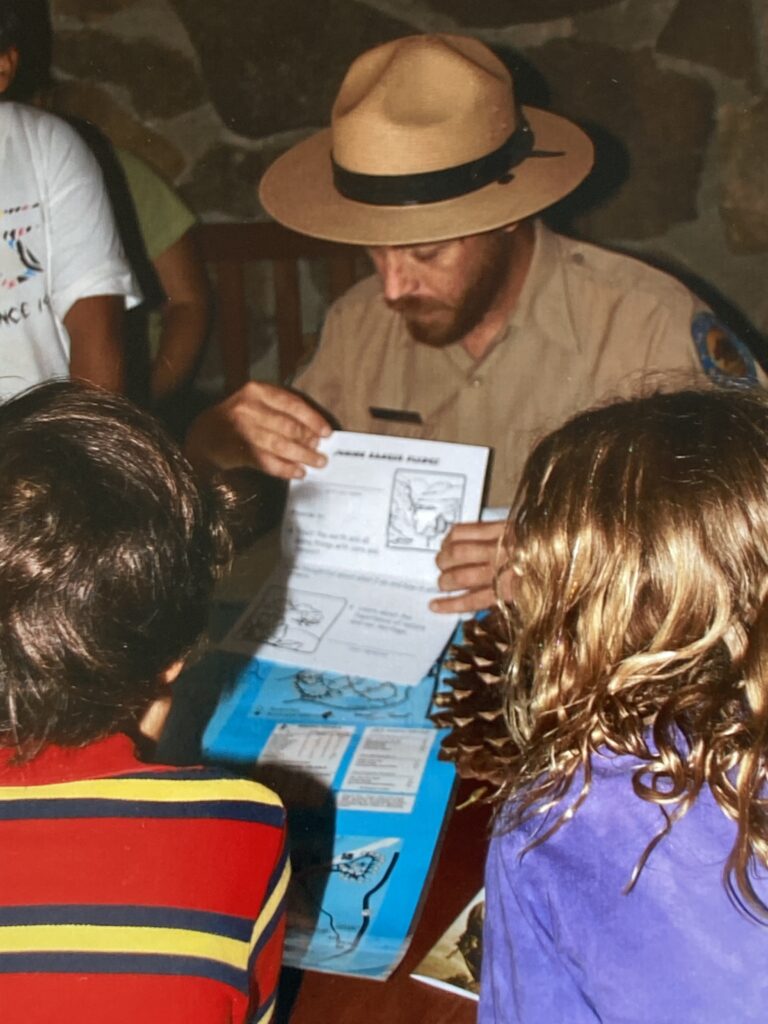 Photograph of a California State Park Interpreter infront of a stone wall and pointing at something in a paper booklet while three youth in the foreground look toward the booklet.