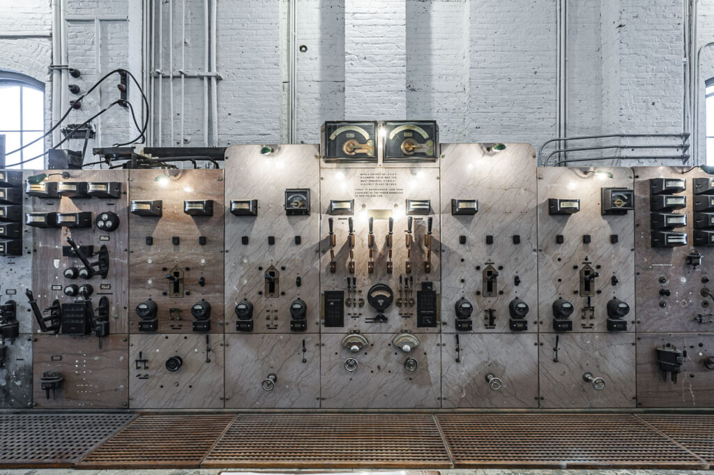 Photograph of an antique electrical powerplant control panel made of marble and featuring knife switches, analog meters, and incandescent lights.