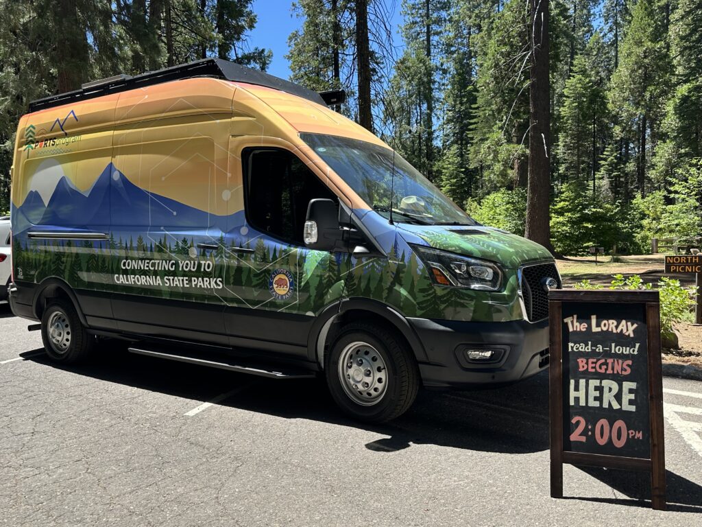 Photograph of a parking lot at Calaveras Big Trees State Park depicting a camper van with a decorative exterior wrap reading 