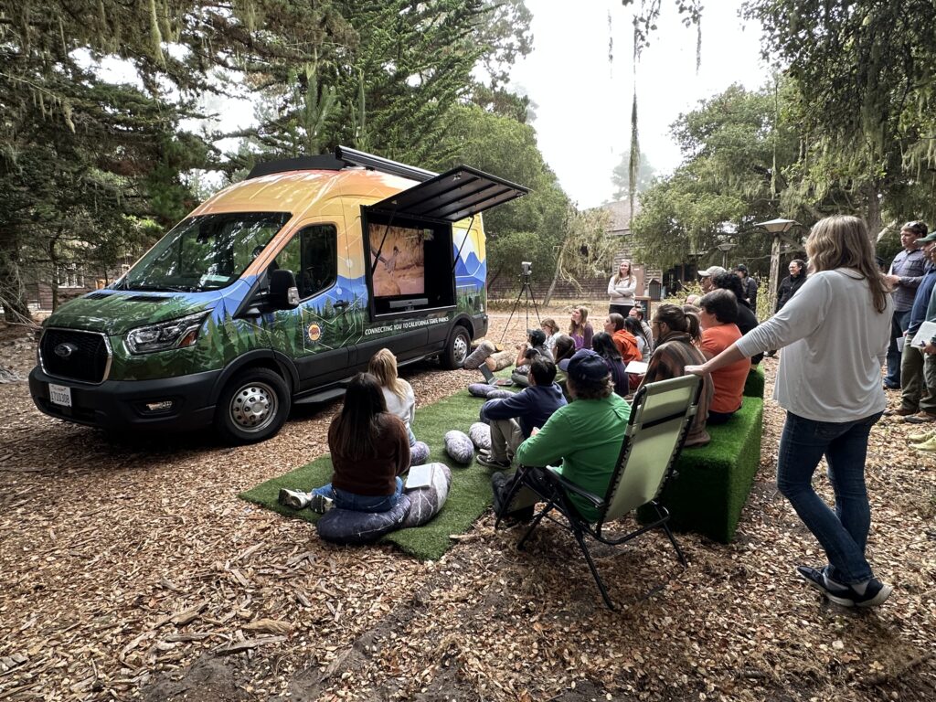 Photograph of a foggy forest of Monterey Cypress trees interspersed with wood shingled buildings depicting the PORTS Van, "VANNIE," with a side panel open revealing a screen; in the foreground a group of adults are standing and seated on chairs and on artificial turn looking at the screen.