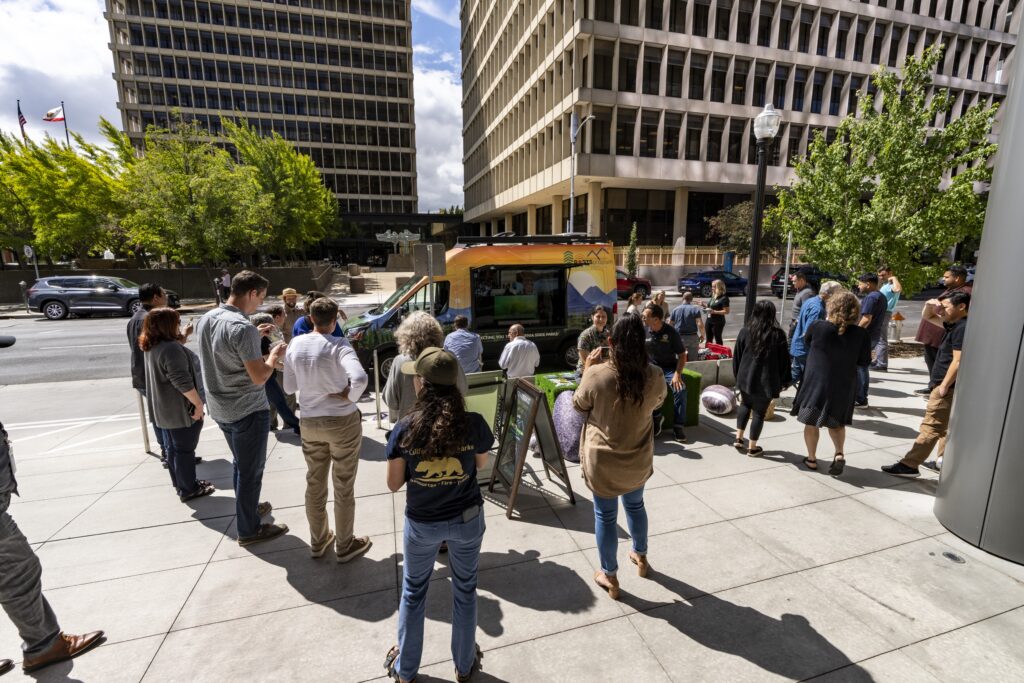 Photograph of the front plaza of the Calfiornia Natural Resources Building in Downtown Sacramento depicting the PORTS Van parked along the street with a side panel open revealing a screen; in the foreground a group of people stand and sit on street furniture looking at the screen.
