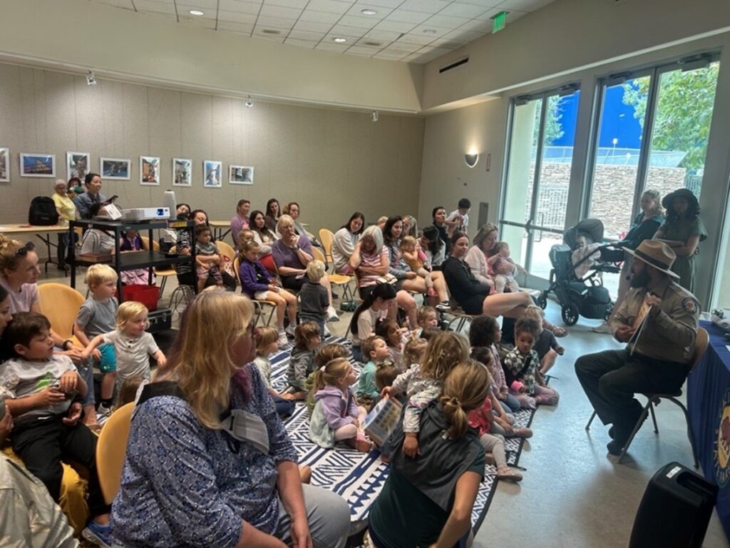 Photograph of the interior of a library event room depicting a seated California State Park Interpreter reading a book to a group of around 30 children and adults variously seated in chairs and on a rug.