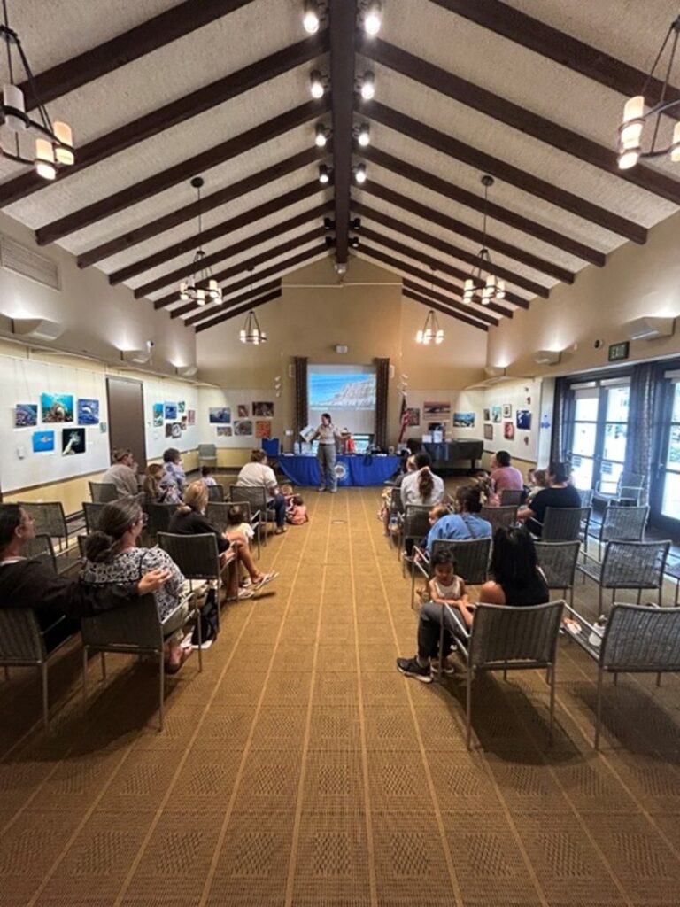 Photograph of a long library event room with vaulted ceilings depiciting a California State Park staff person standing infront of a screen and a community outreach table reading to a group of 15 adults and children seated in chairs.