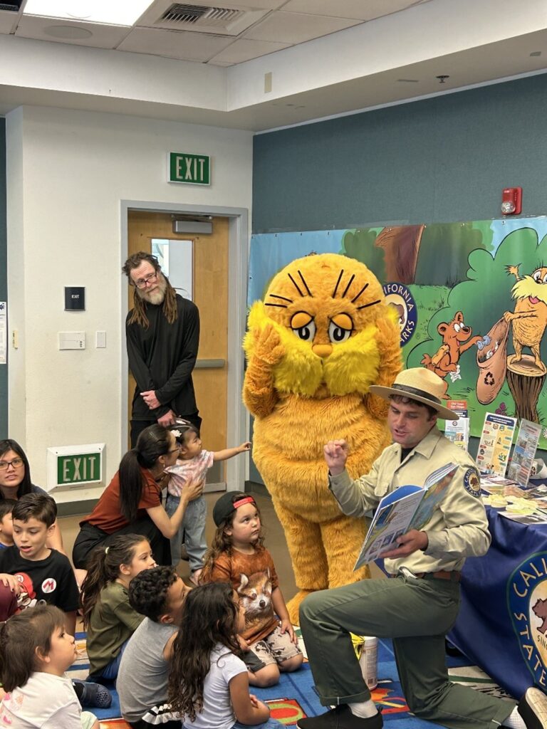 Photograph of the interior of a library depicting a California State Park Interpreter kneeling in front of a community outreach table while reading a book to a group of children seated in front of him, while behind him stands an adult and a person in a Lorax mascot costume.