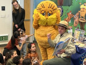 Photograph of the interior of a library depicting a California State Park Interpreter kneeling in front of a community outreach table while reading a book to a group of children seated in front of him, while behind him stands an adult and a person in a Lorax mascot costume.