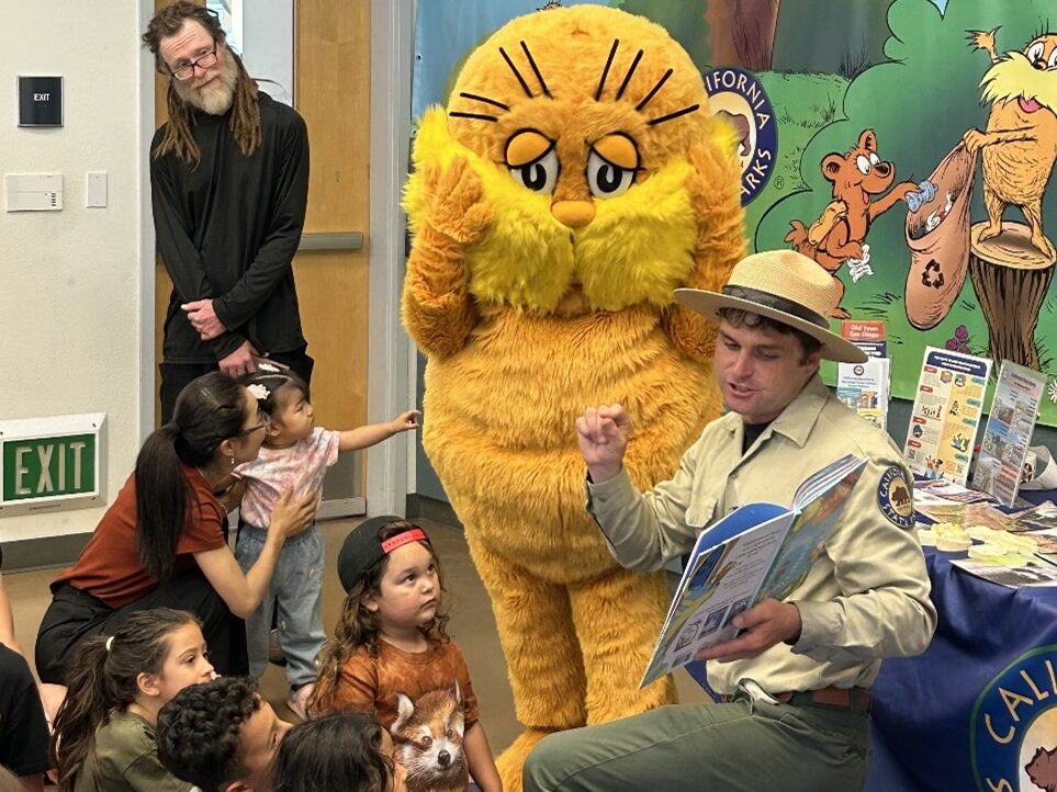 Read with a Ranger Photograph of the interior of a library depicting a California State Park Interpreter kneeling in front of a community outreach table while reading a book to a group of children seated in front of him, while behind him stands an adult and a person in a Lorax mascot costume.