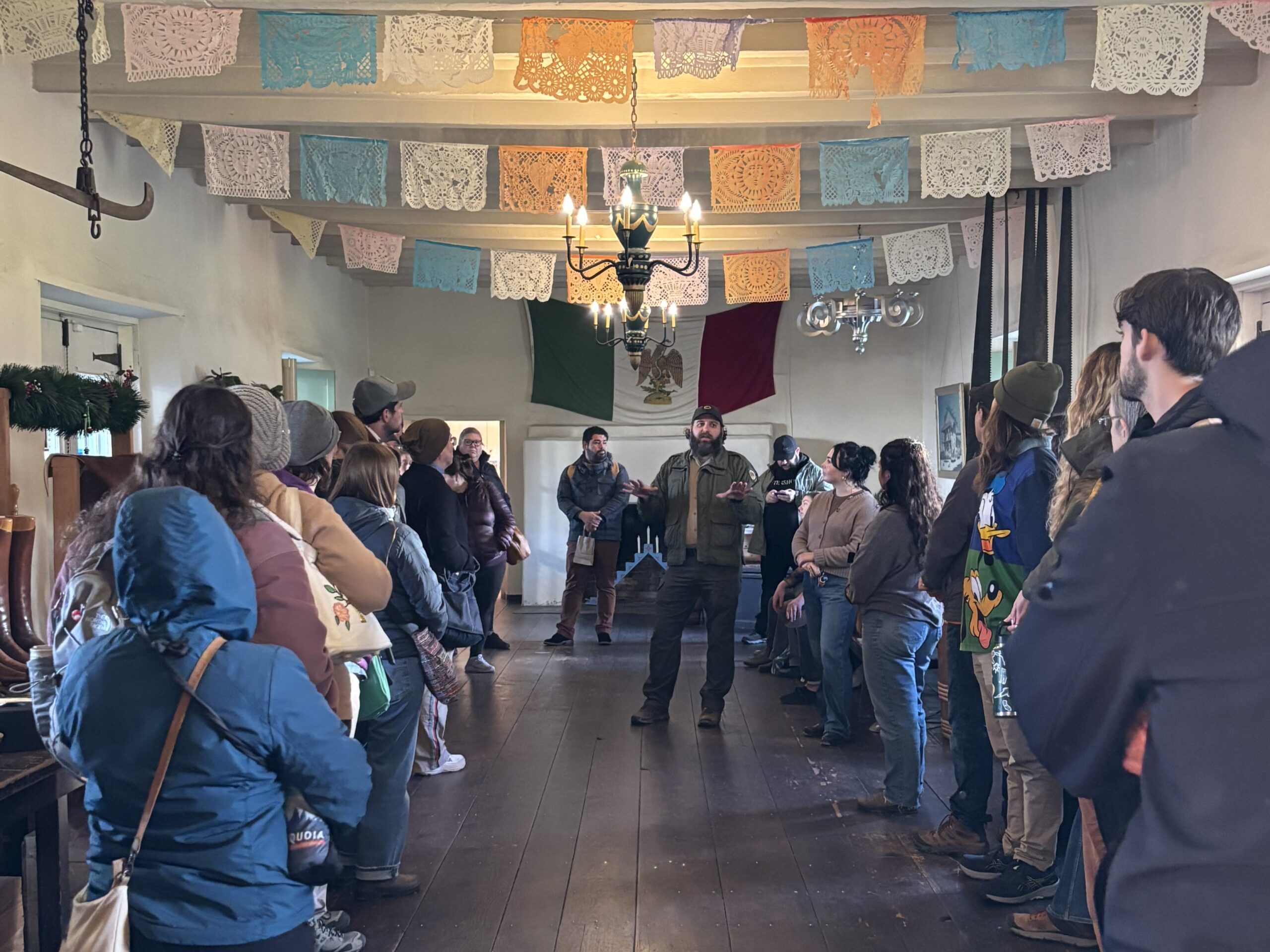 Photograph looking down the interior of a long, narrow room with adobe walls, tile floor, and exposed wood rafters, decorated with a candle chandelier, papel picado banners, and a historic era Mexican flag; the room is populated with adults lined up along either facing wall with their backs to the camera and looking towards a person in California State Parks uniform facing the camera and delivering a talk.