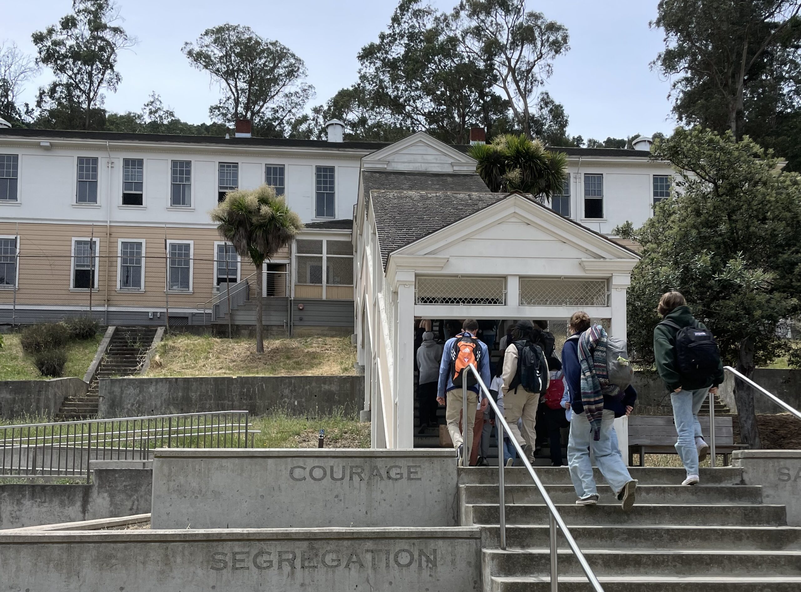 A group of students walk up concrete steps toward a historic white barracks building at Angel Island State Park, surrounded by trees and hillside vegetation.
