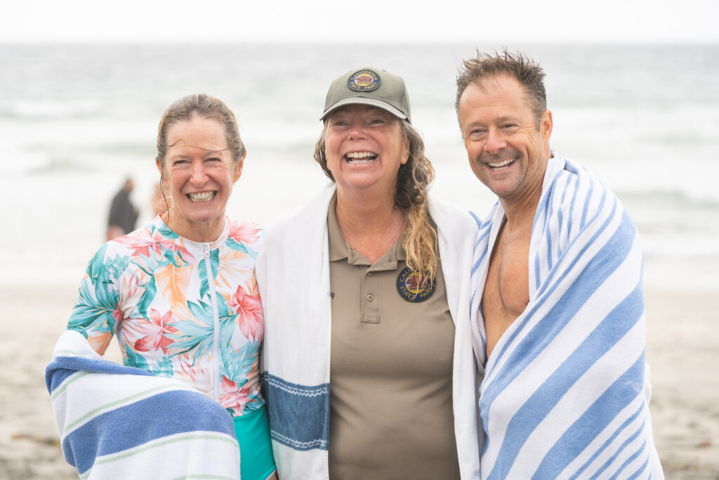 Photograph of three wet and smiling people--two dressed in towels and swimwear, and one wearing a California State Parks ballcap and polo--standing arm in arm on a beach.