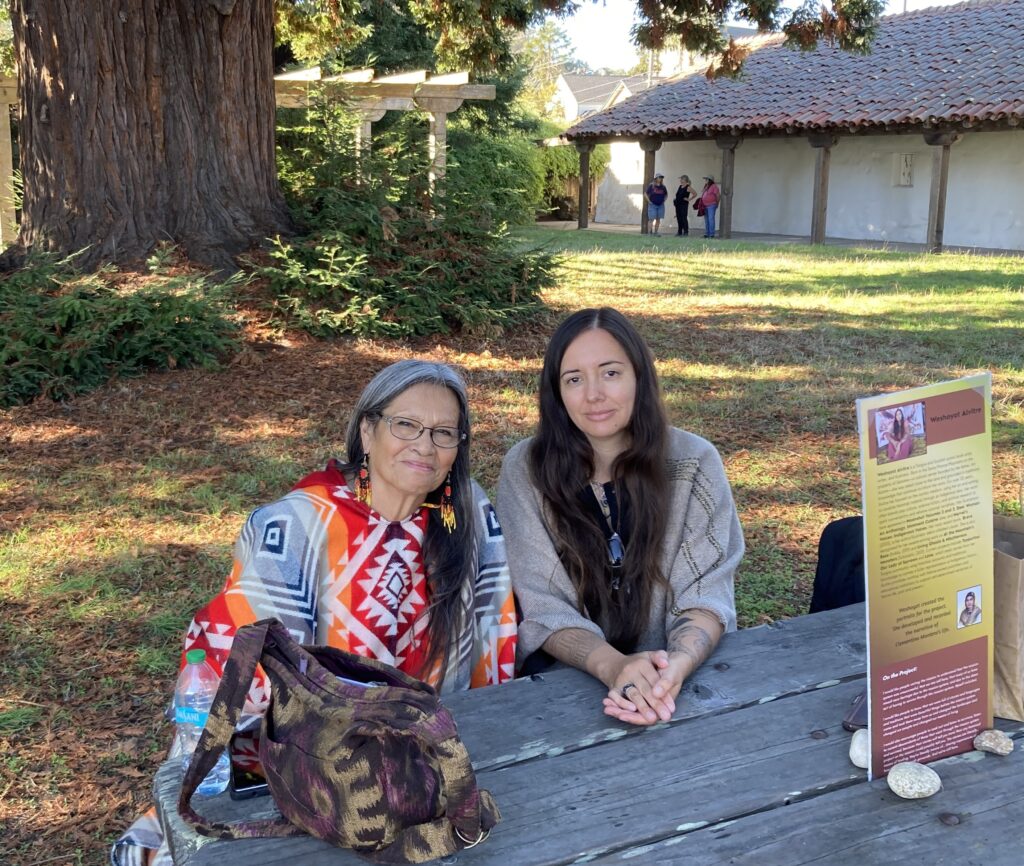 Photograph of two people seated at a picnic table outdoors with a redwood tree and an adobe walled and tiled roof structure behind them.