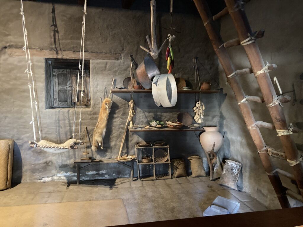 Photograph of the interior of a museum room at Mission Santa Cruz State Historic Park featuring adobe walls and floors and furnished with everyday living items of Native People at the Mission, many of which blend Native Californian items and forms with Spanish Colonial items and forms.