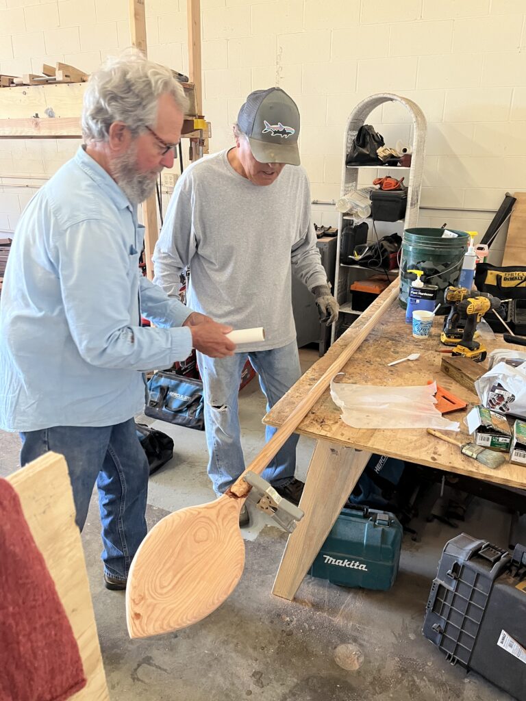 Photograph of an interior work space featuring two men applying a binding to a wooden panel lad out on a work table in front of them, strewn with various tools.