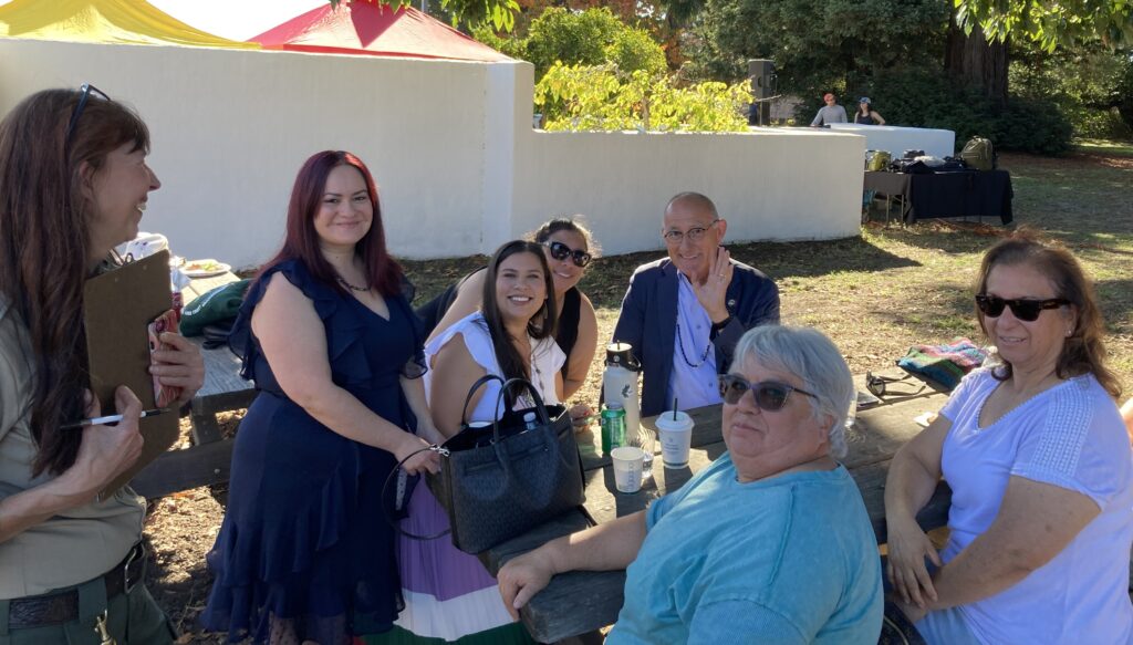 Photograph of a group of seven people seated and standing around a picnic table outdoors with a low white wall behind them.