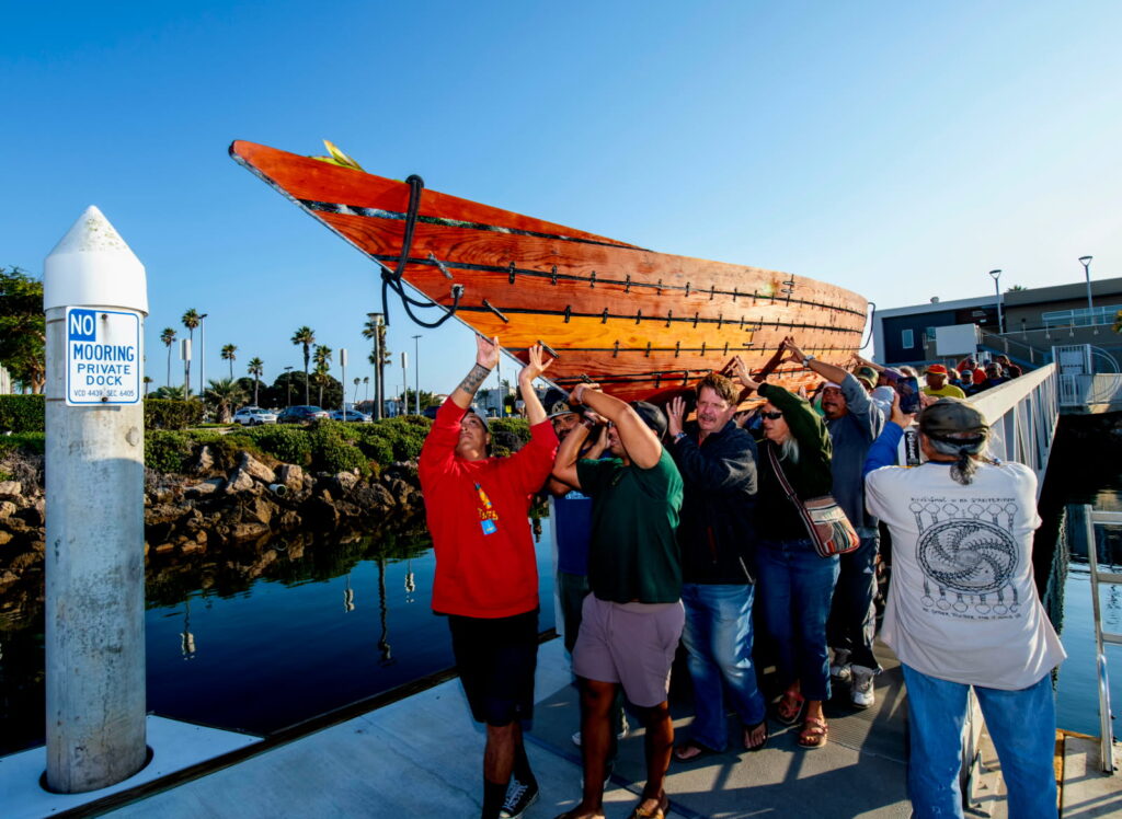 Photograph of a marina featuring a dozen people walking down a dock hoisting a wooden sewn-plank Chumash tomol canoe over their head, while a dozen more people walk behind them.